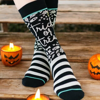 A pair of women's Halloween socks with black and gray stripes and 'Trick or Treat' text pattern, displayed on a wooden surface with lit candles and pumpkins around.