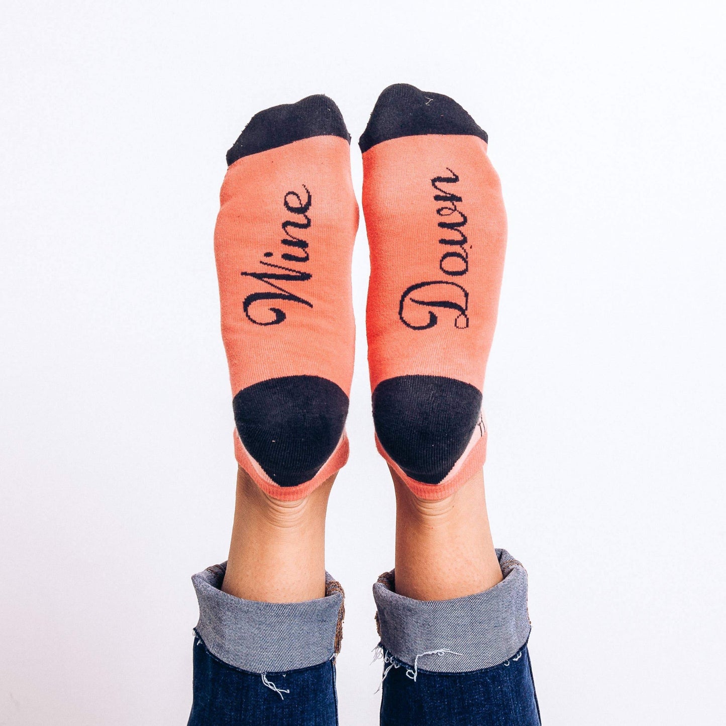 Coral-colored ankle socks with navy blue heels and toes, decorated with a pattern of wine glasses and wine bottles and "Wine Down" written on the soles. Great women's wine gift