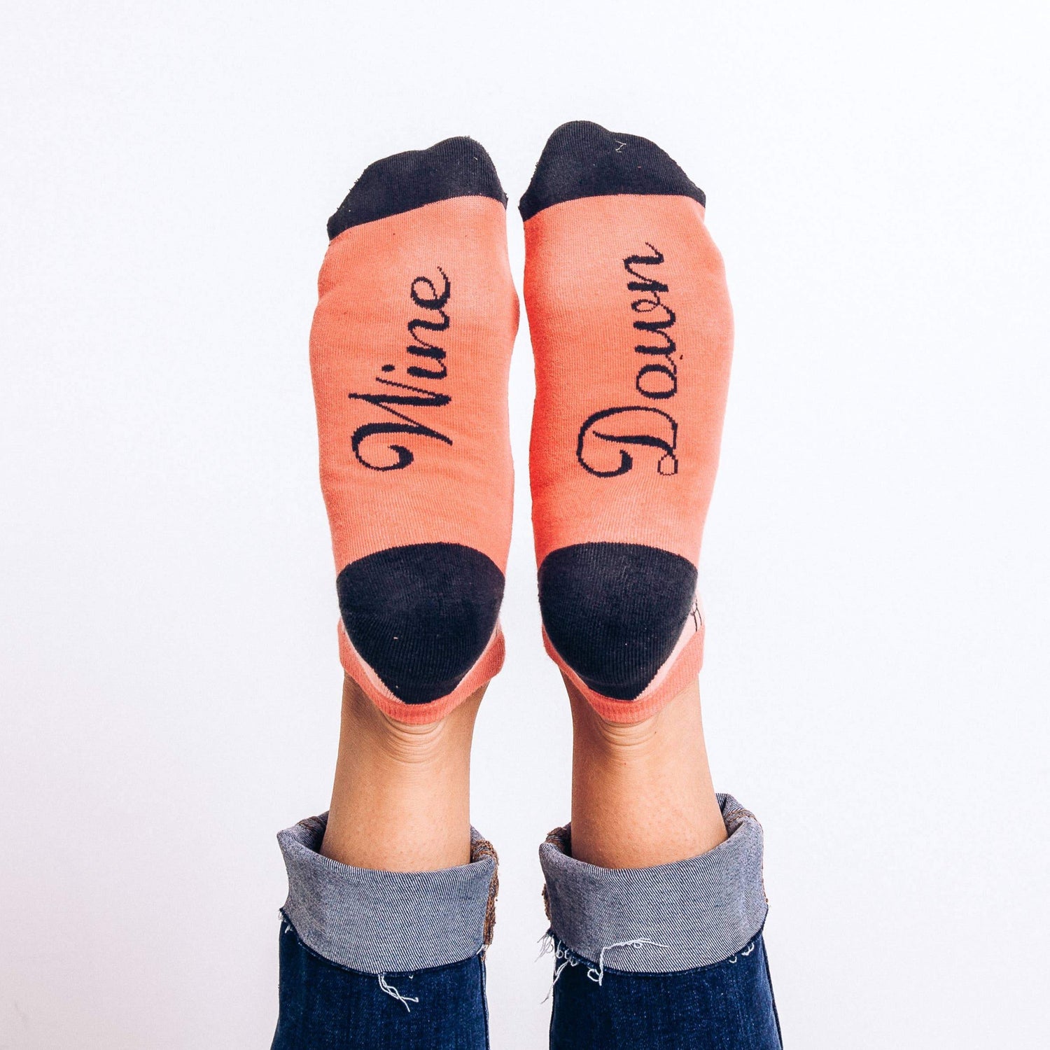 Coral-colored ankle socks with navy blue heels and toes, decorated with a pattern of wine glasses and wine bottles and "Wine Down" written on the soles. Great women's wine gift