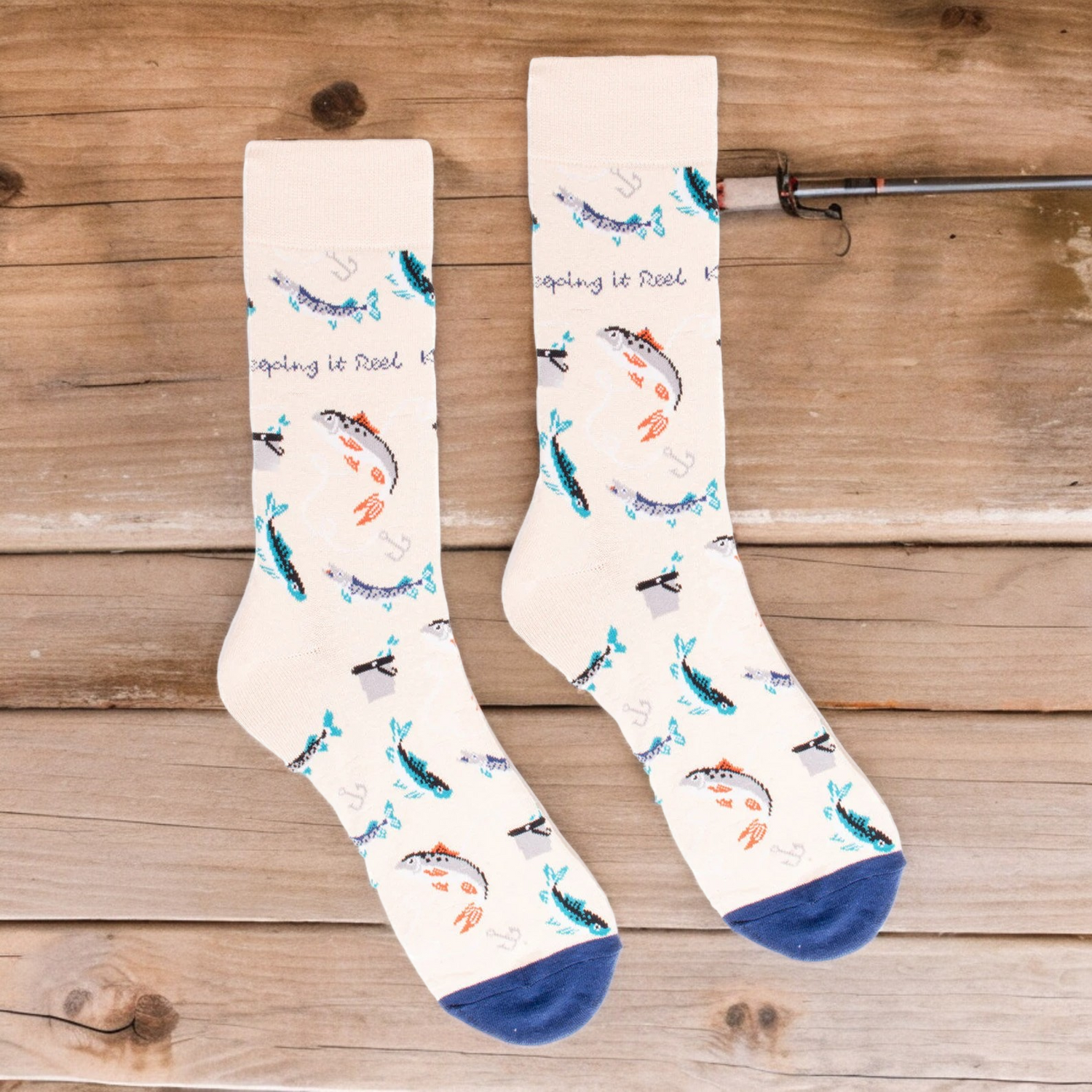A pair of cream-colored men's socks featuring a playful fishing theme. The socks are decorated with various fish, fishing hooks, and anchors in shades of blue, teal, and orange. The phrase "Keeping it Reel" is scattered throughout the design. The toes are accented with a navy blue color.
