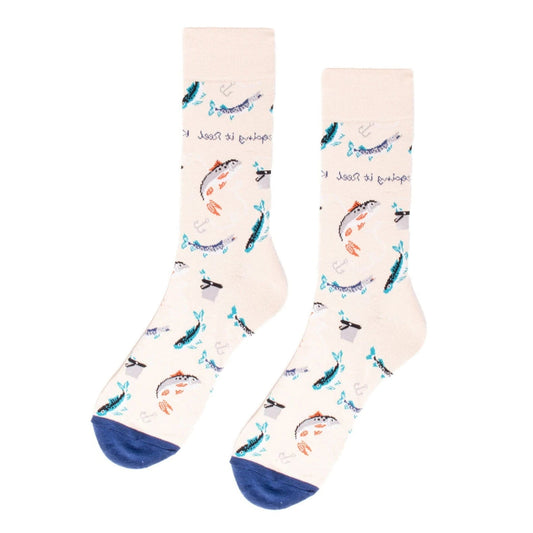 A pair of cream-colored men's socks featuring a playful fishing theme. The socks are decorated with various fish, fishing hooks, and anchors in shades of blue, teal, and orange. The phrase "Keeping it Reel" is scattered throughout the design. The toes are accented with a navy blue color.
