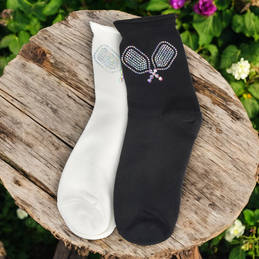 A pair of women's socks, one in black and one in white, both with a rhinestone tennis racquet design on the ankle area, displayed on a wooden surface with flowers in the background.